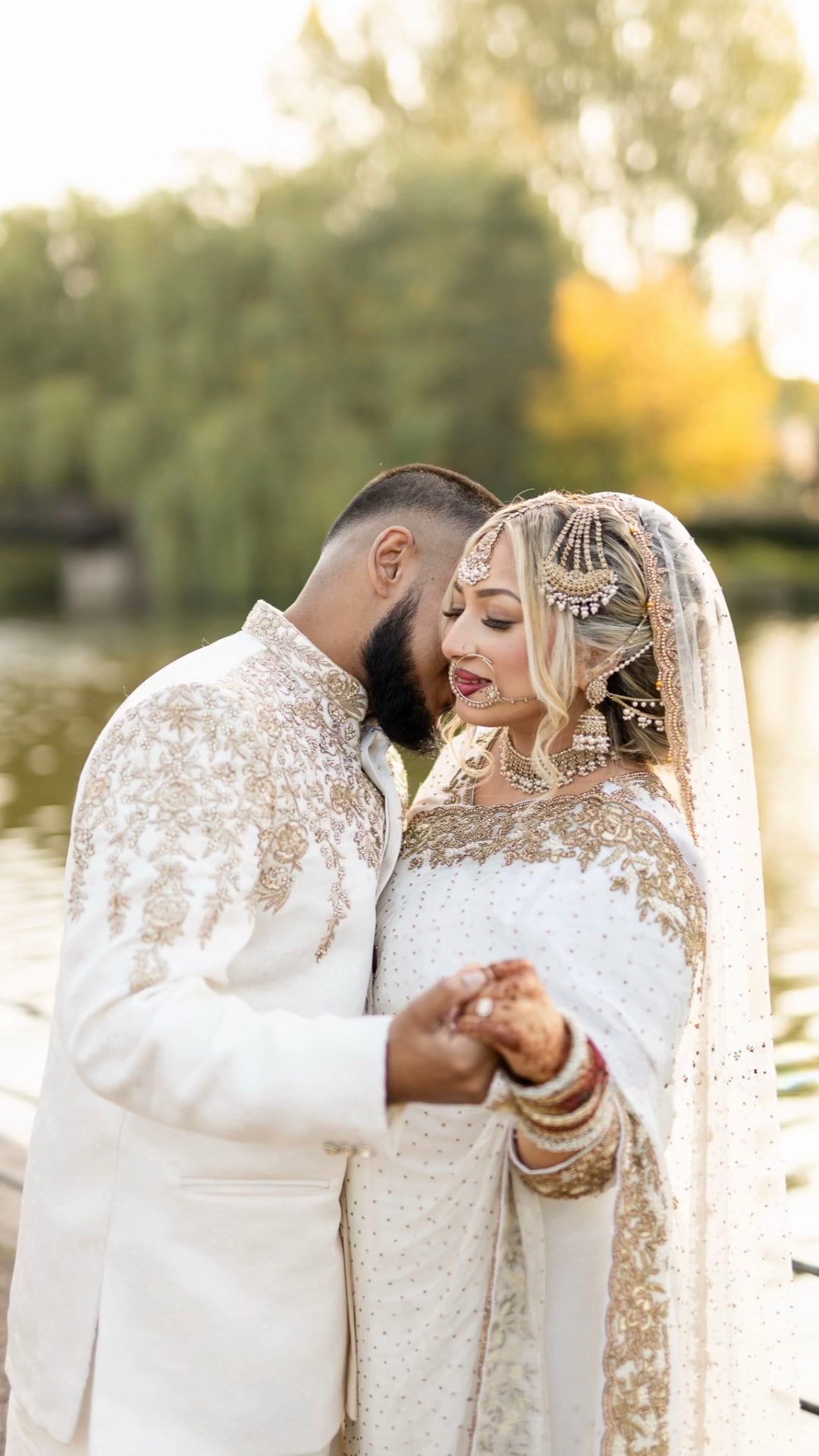 Couple in ivory and gold holding hands beside a lake in autumn light