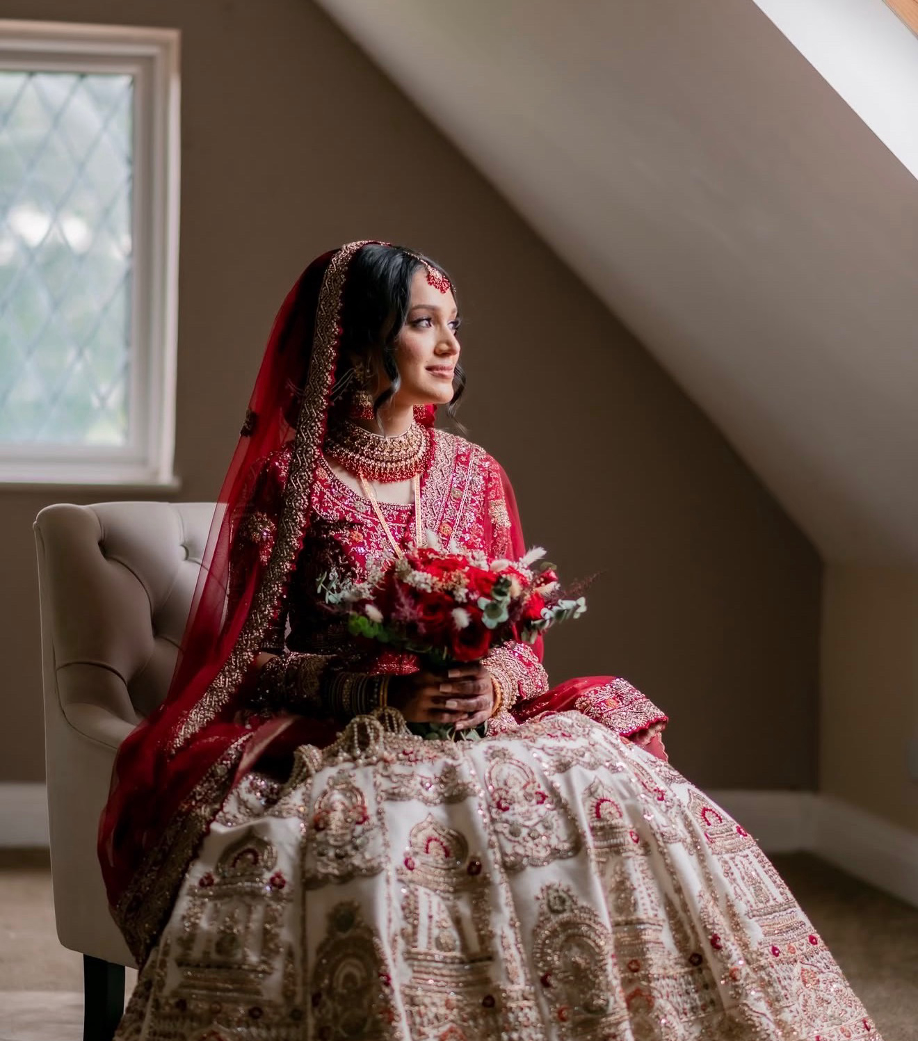 Bride in red bridal wear seated by a window holding a red and white bouquet