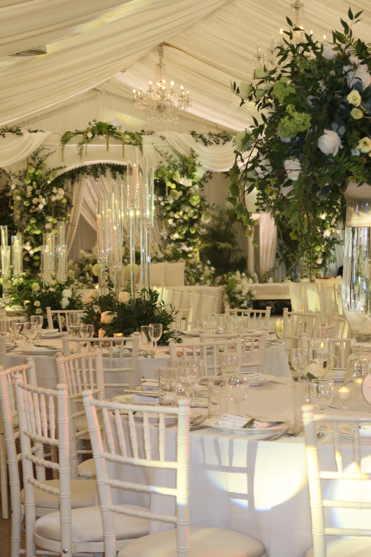 Marquee laid with white chiavari chairs, glassware and tall green and white florals