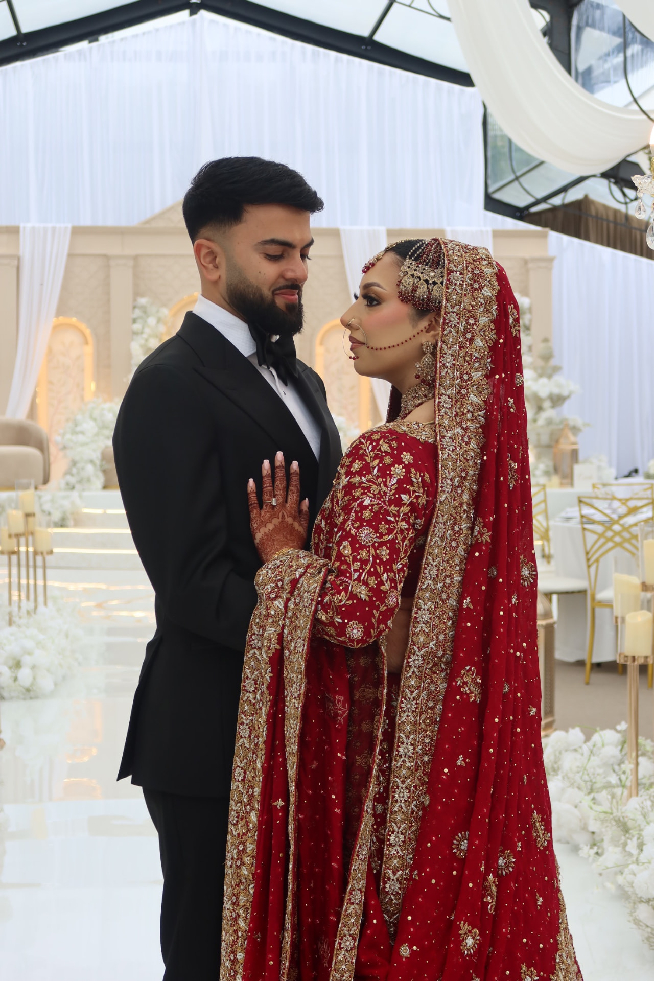 Bride in a red and gold lengha standing with the groom in a black tuxedo inside a draped marquee