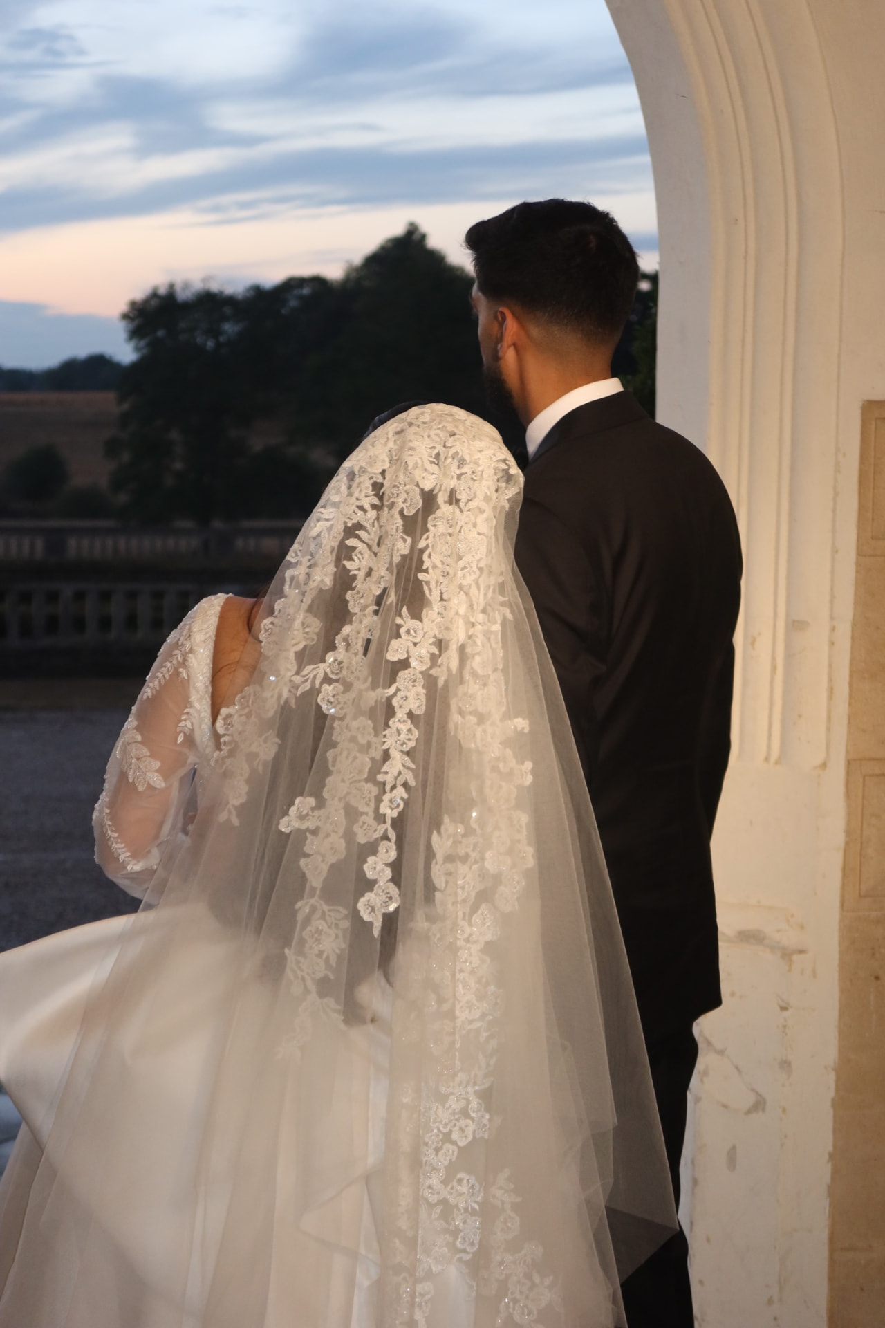 Couple seen from behind under a stone arch at dusk, the bride in a lace veil