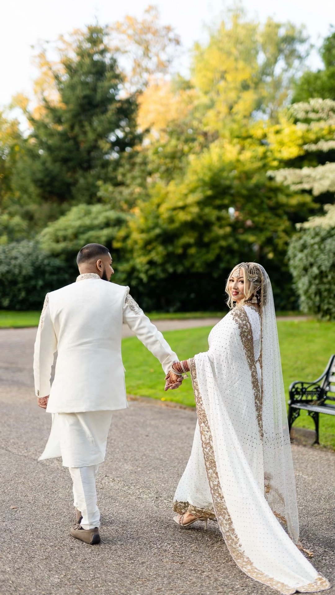 Couple in ivory and gold walking hand in hand along a garden path, the bride looking back