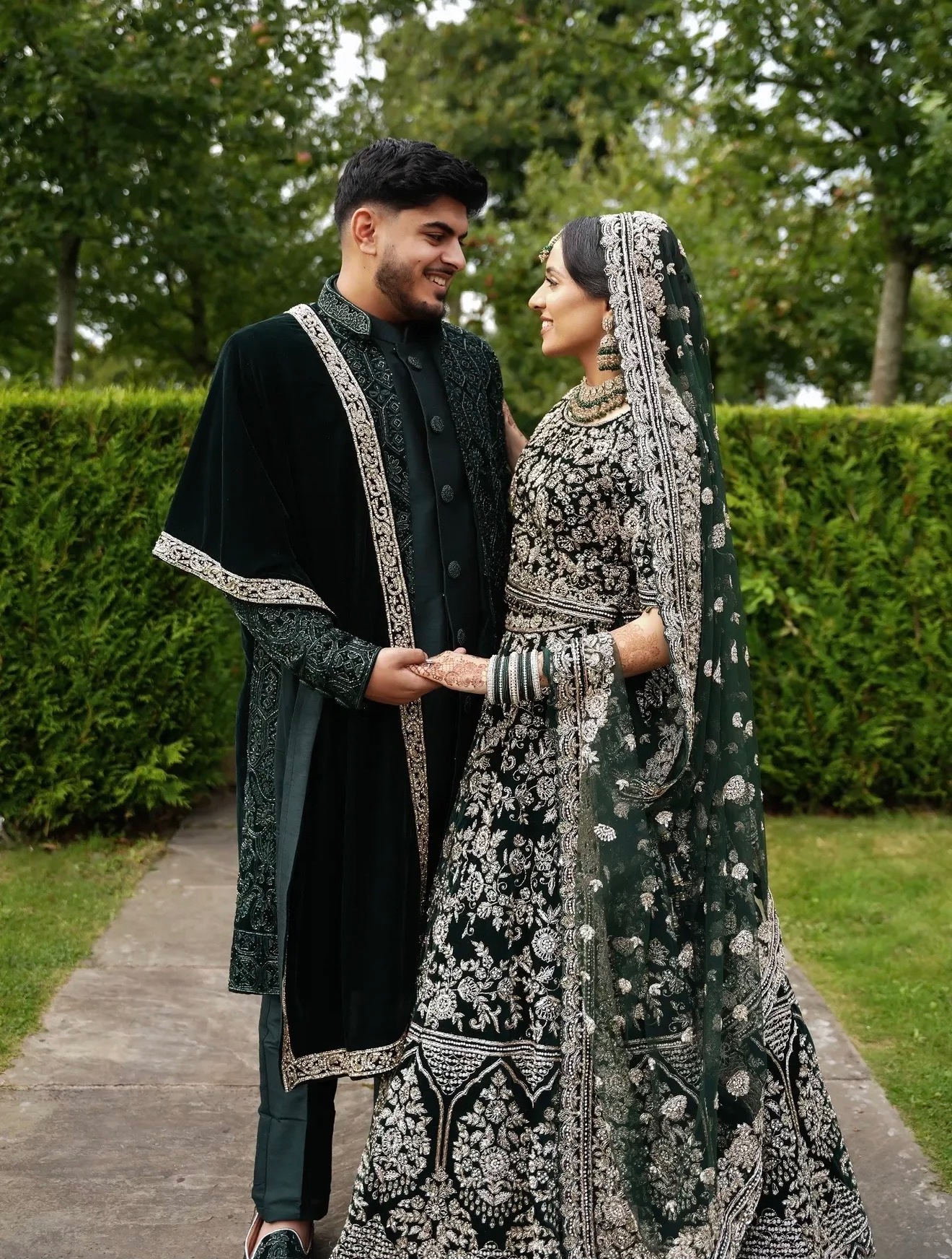 Couple in deep green and silver outfits holding hands on a garden path