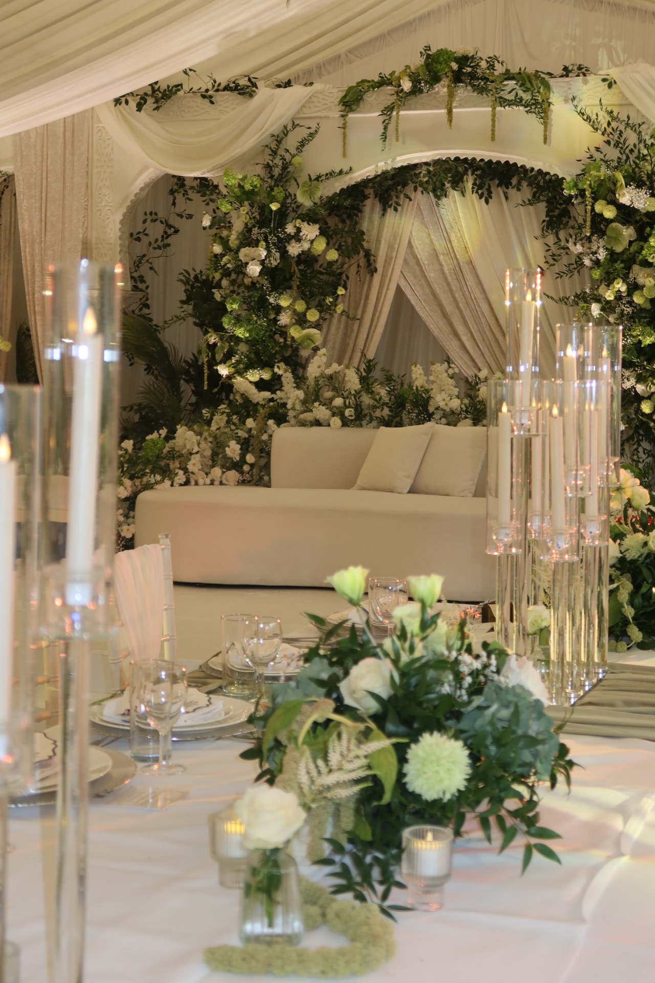 Ivory and green reception stage with tall candles, foliage and a cream sofa
